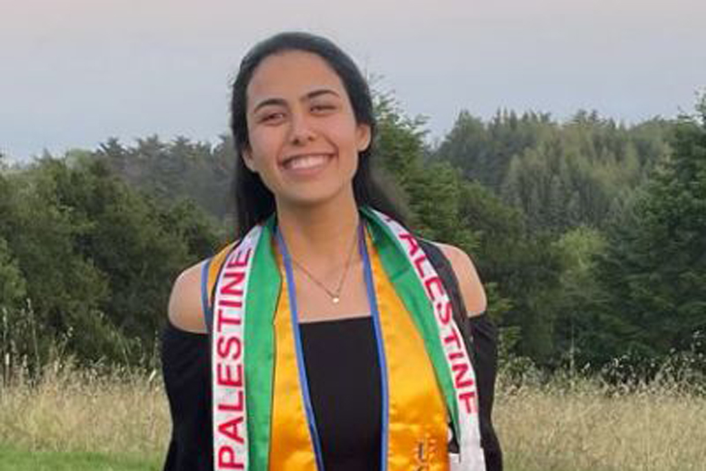 Nabeela Ariqat smiles at the camera. She wears a black dress and a yellow UC Santa Cruz Class of 2021 stole and a Palestinian flag stole. Trees and grass fields are visible in the background.