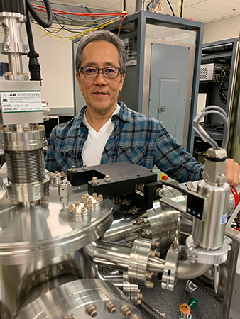 Nobby Kobayashi smiles while standing behind silver lab equipment