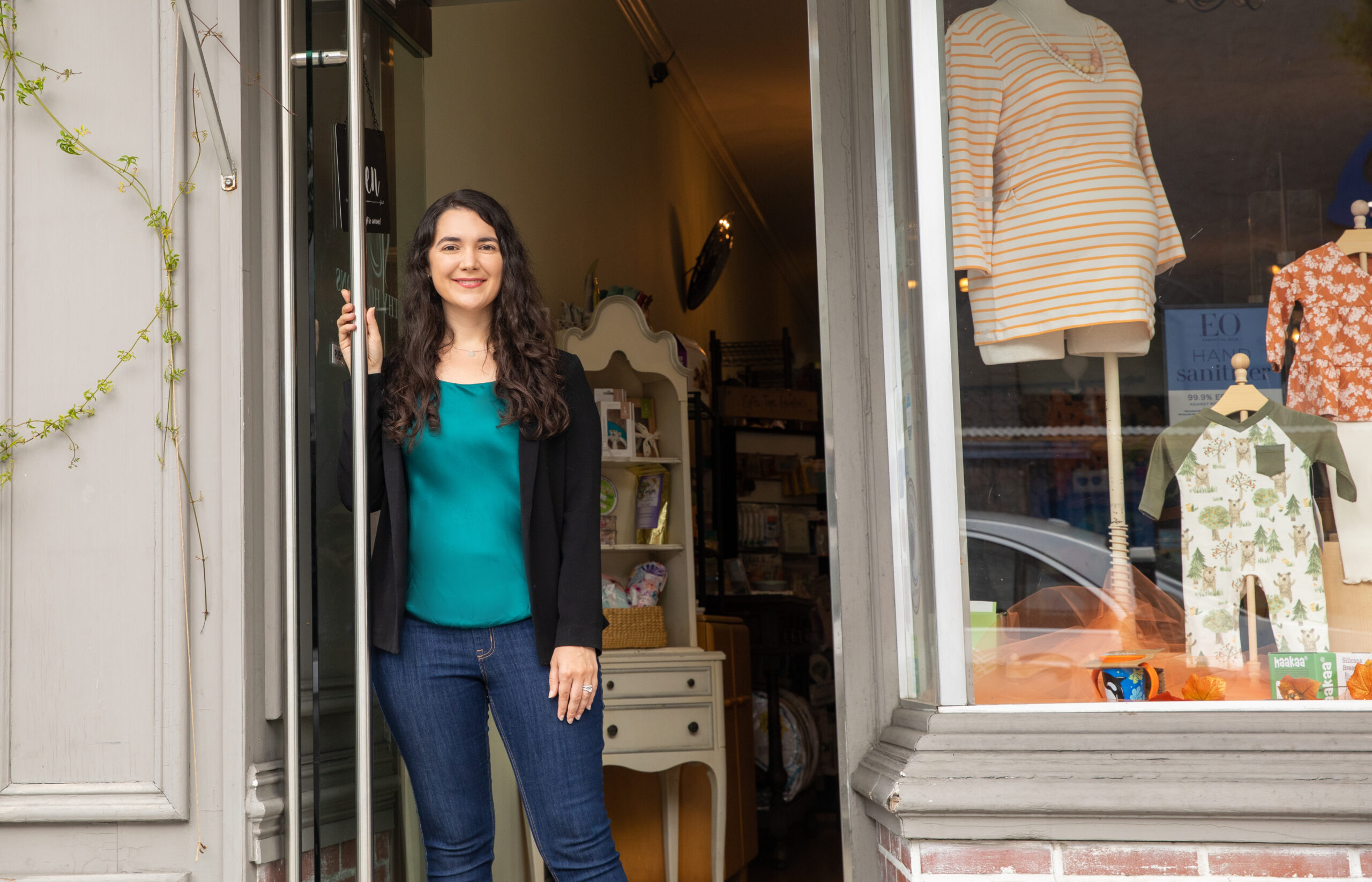 Cassi Janakos stands in the doorway of Peninsula Breastfeeding shop. She is wearing a teal top and black jacket wtih dark denim jeans. She is holding the door open with her right hand