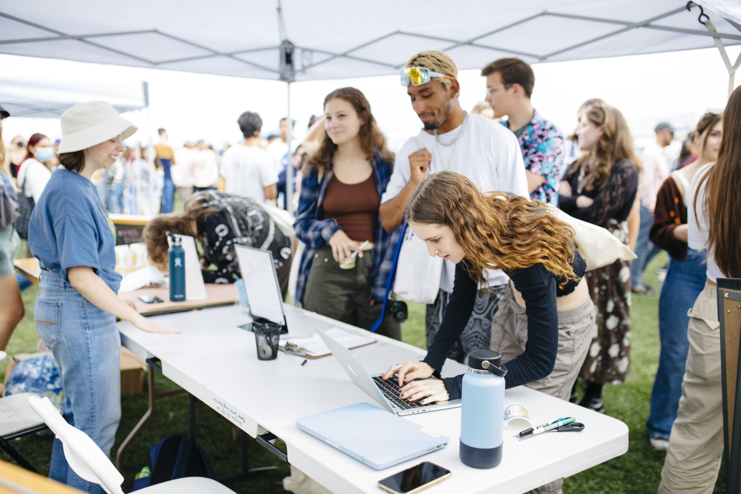 Students gather at an outdoor campus fair, engaging with booths under pop-up tents. One student types on a laptop while others wait in line and chat nearby.