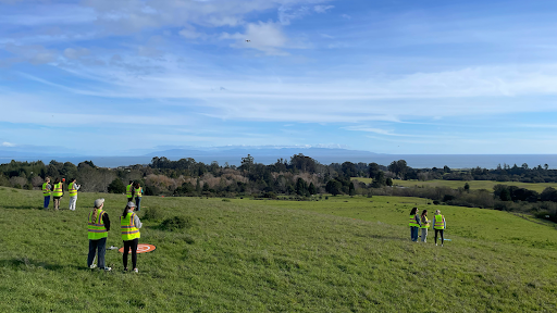 Groups of people wearing yellow safety vests stand in an open grassy field under a blue sky, participating in a drone training program overlooking the distant coastline and ocean.