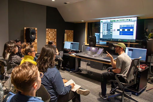 A group of UC Santa Cruz graduate students sit facing a large mixing console and multiple monitors in a professional audio studio, listening to an instructor who gestures toward a projection of audio editing software during a workshop in the new Social Documentation Laboratory.