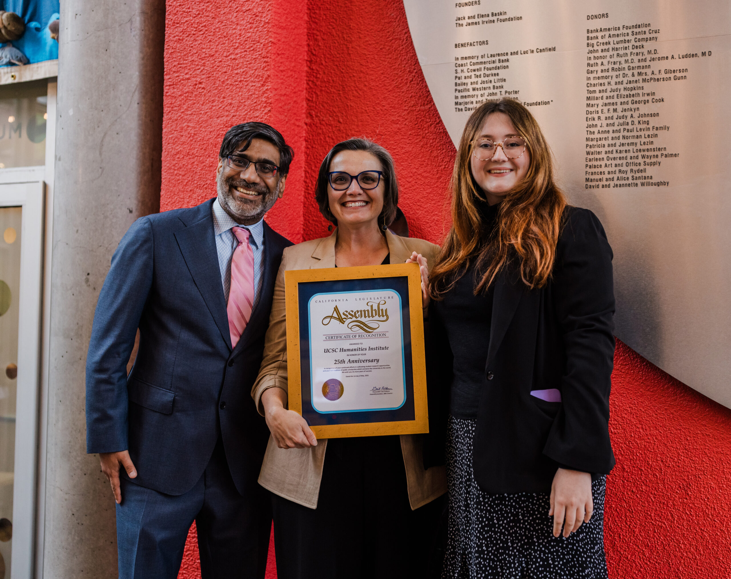 three people stand in front of a red background, smiling and holding up a large plaque.