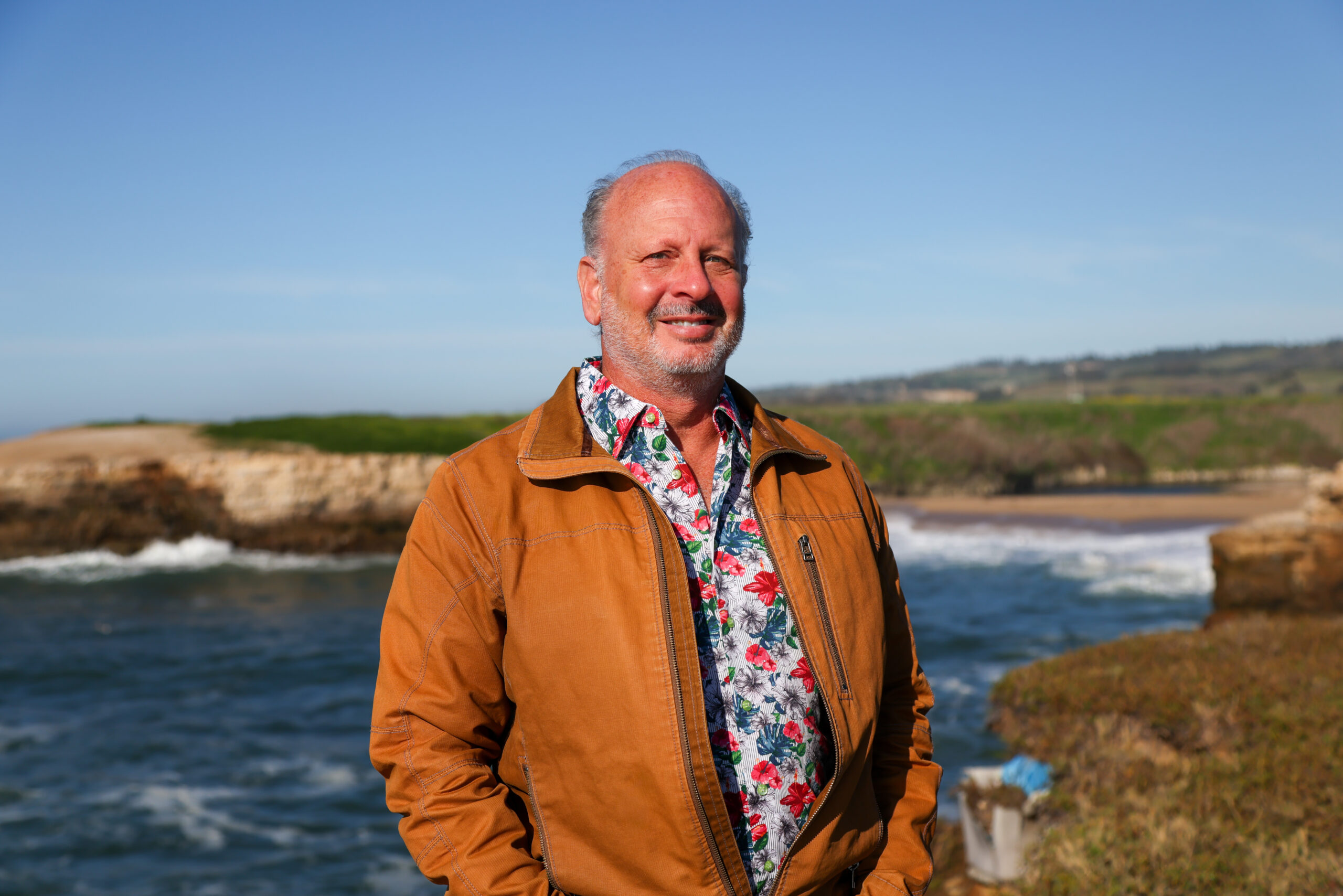 Mike Beck in a floral shirt and rust jacket by the Monterey Bay coastline.