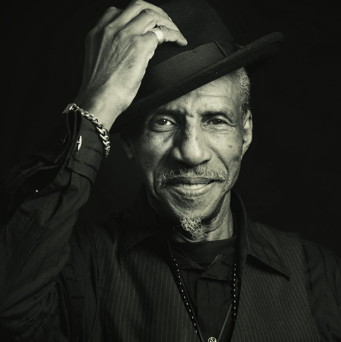 An older man wearing a dark hat and shirt smiles warmly while tilting his hat. He sports a goatee, a chain bracelet, and a beaded necklace.