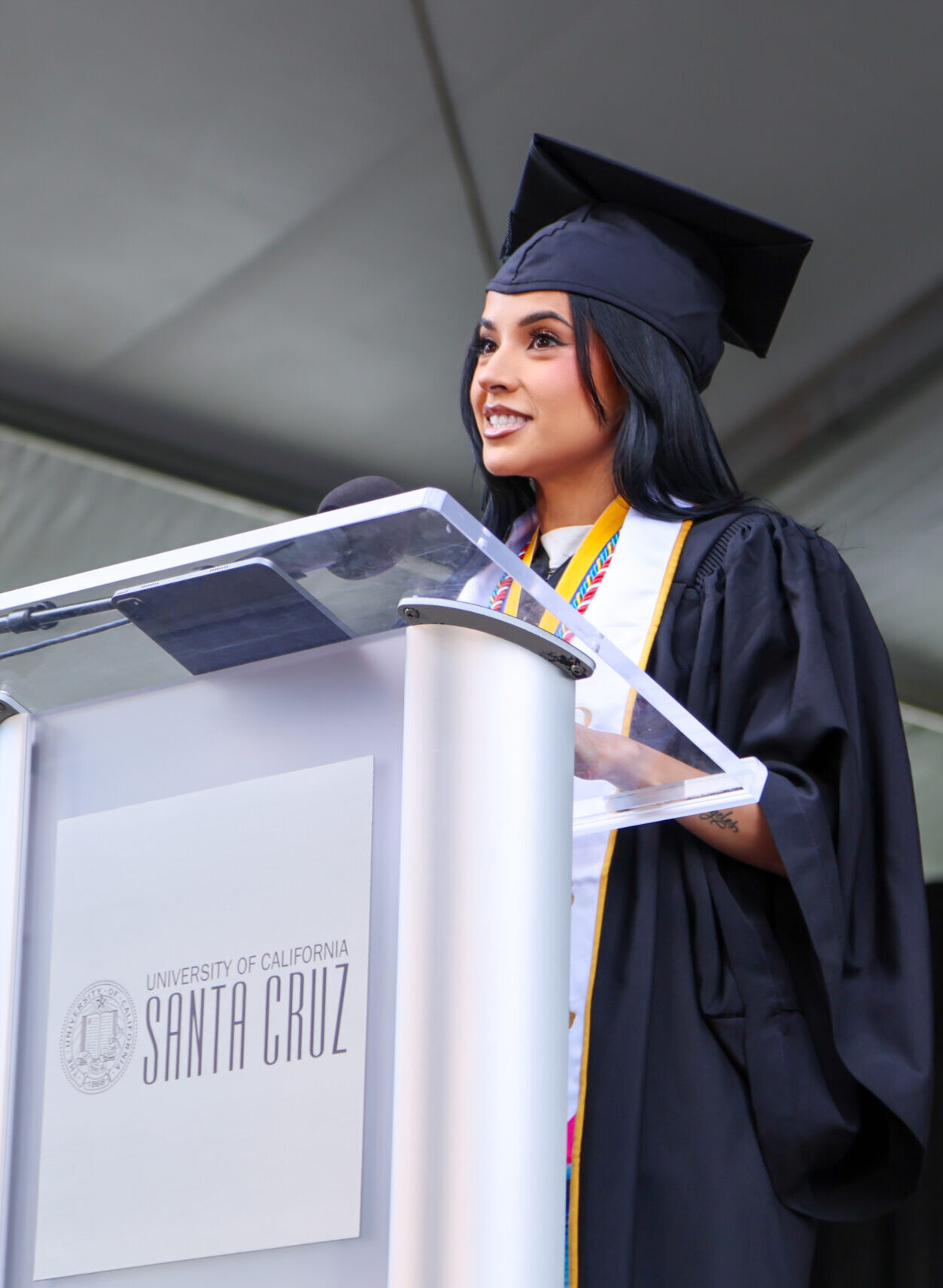 Becky G in cap and gown speaks at a podium under a large tent. She wears a medal and stole while delivering her speech.