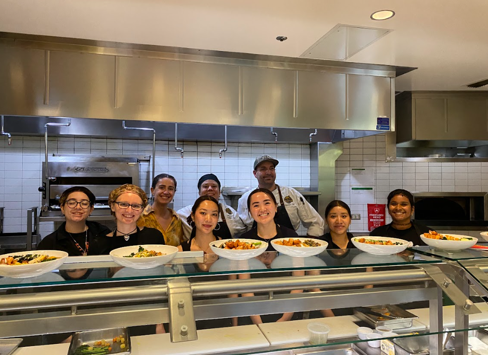 A group of UC Santa Cruz students and dining staff stand together behind a counter in a university kitchen, smiling behind row of prepared healthy bowls.