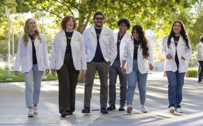 Six smiling students in white lab coats walk outdoors under green trees. They appear relaxed and cheerful.
