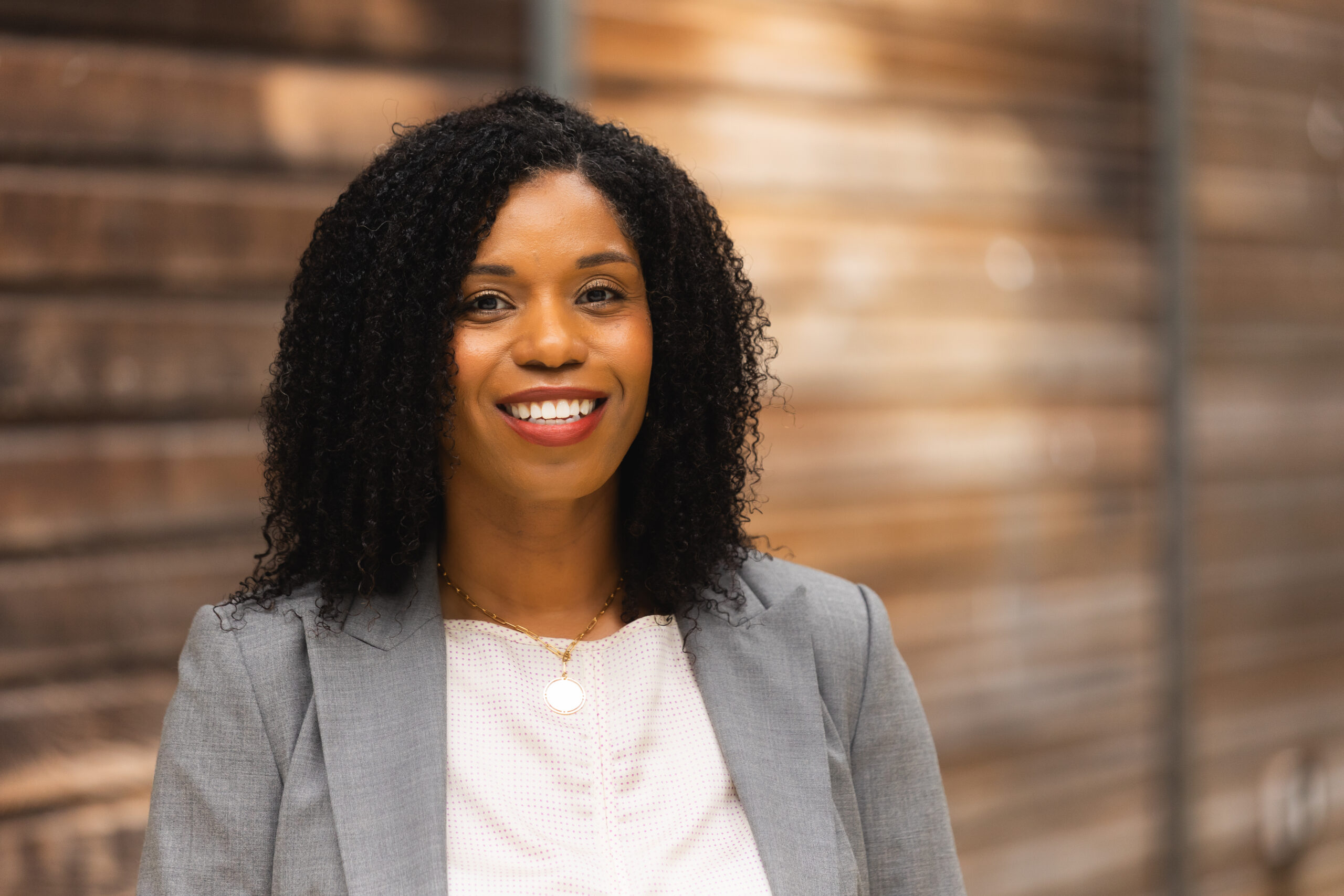 A woman with curly hair smiles while wearing a gray blazer and white top. The background features blurred wooden panels.