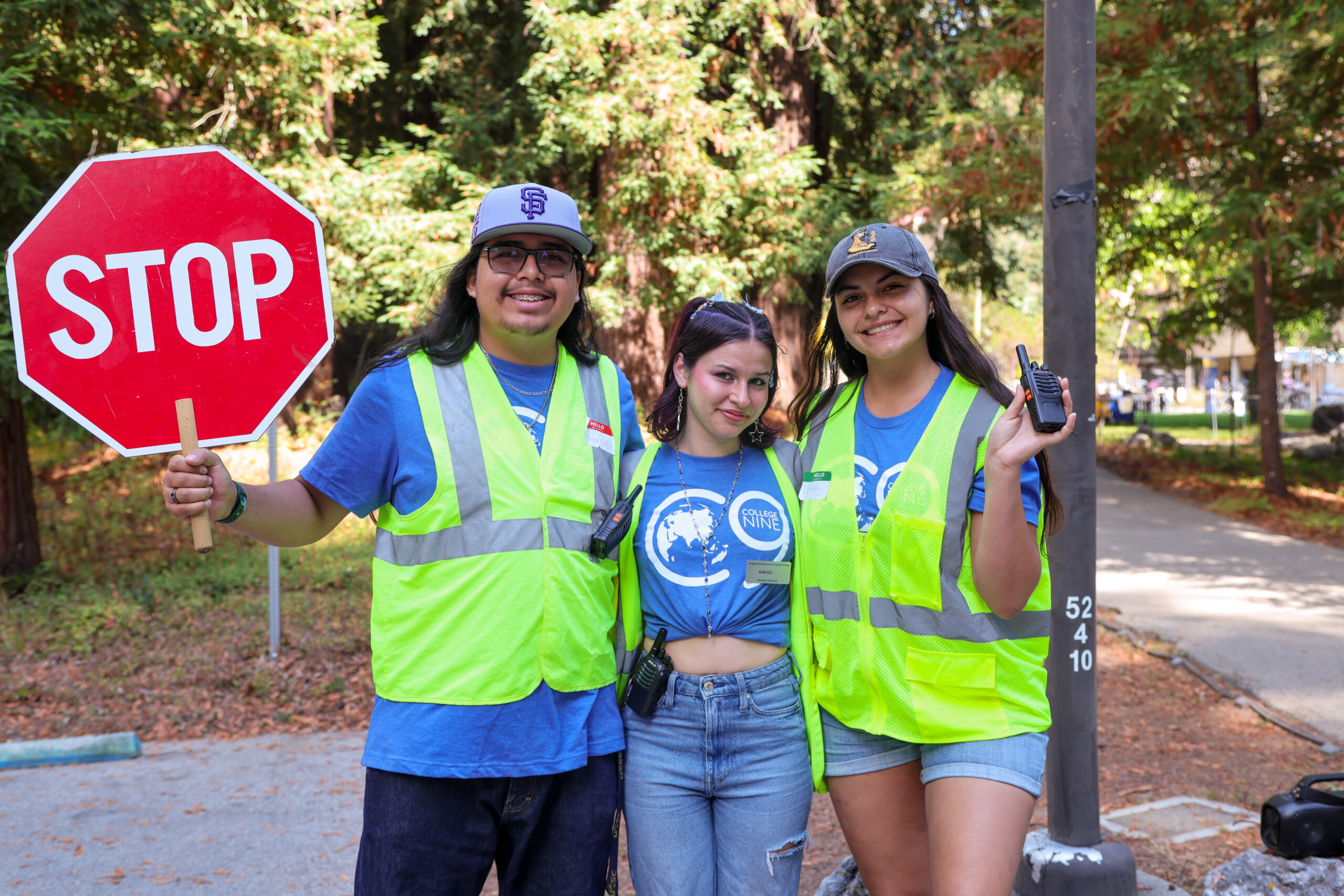 Three student move-in volunteers wearing blue shirts and bright safety vests pose together outdoors. One holds a stop sign and another holds a walkie-talkie.