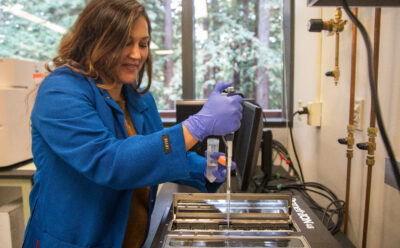 A woman wearing a blue lab coat pipettes into a large DNA sequencing device.