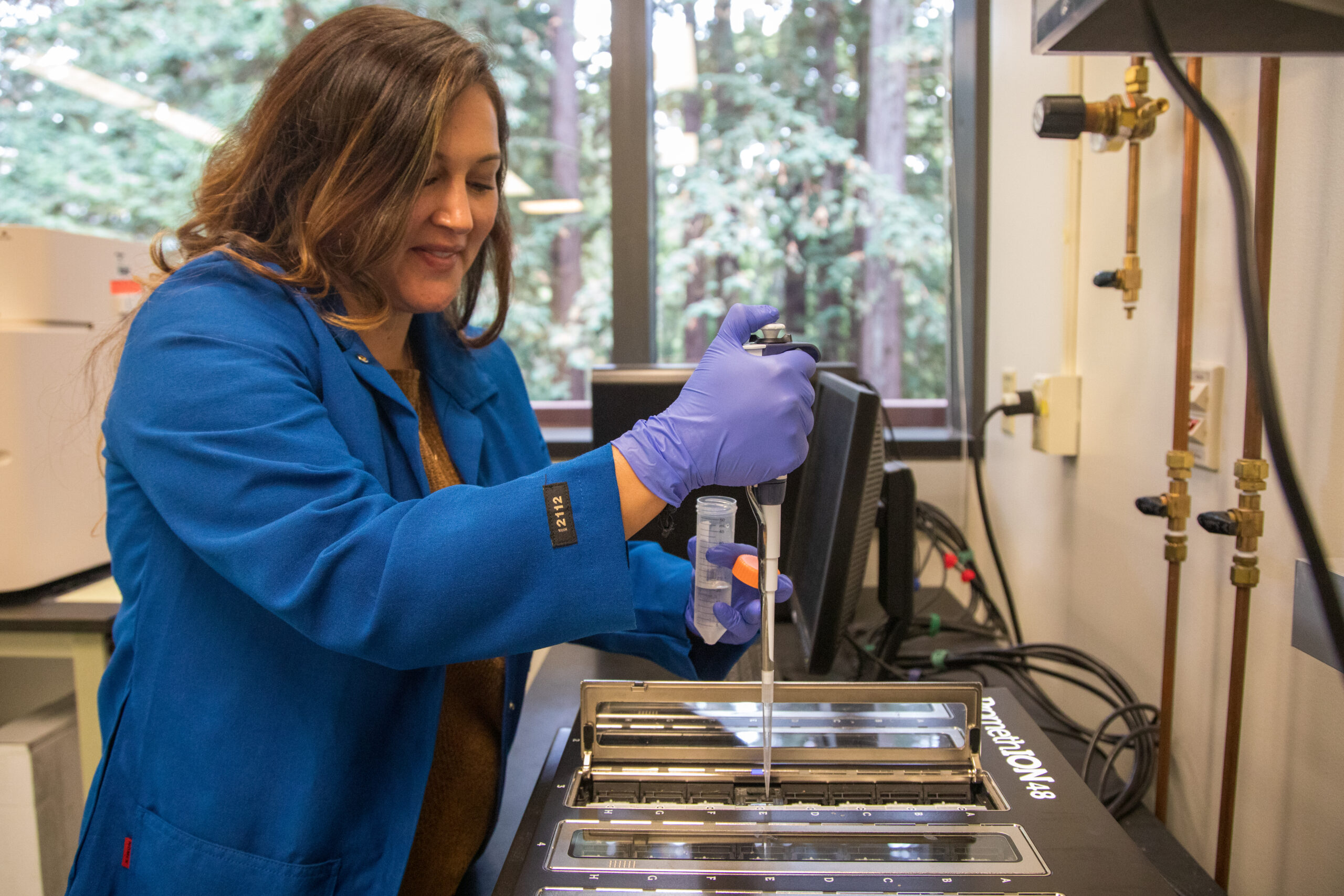 A woman wearing a blue lab coat pipettes into a large DNA sequencing device.