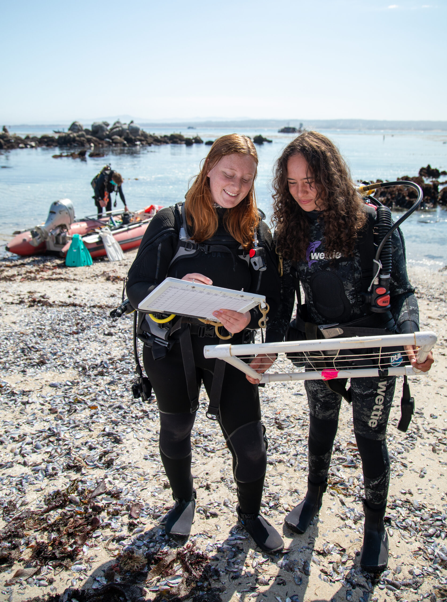 Two scuba divers in wetsuits stand on a rocky beach, one holding a clipboard and the other a survey frame. A small inflatable boat and another diver are visible near the shoreline.