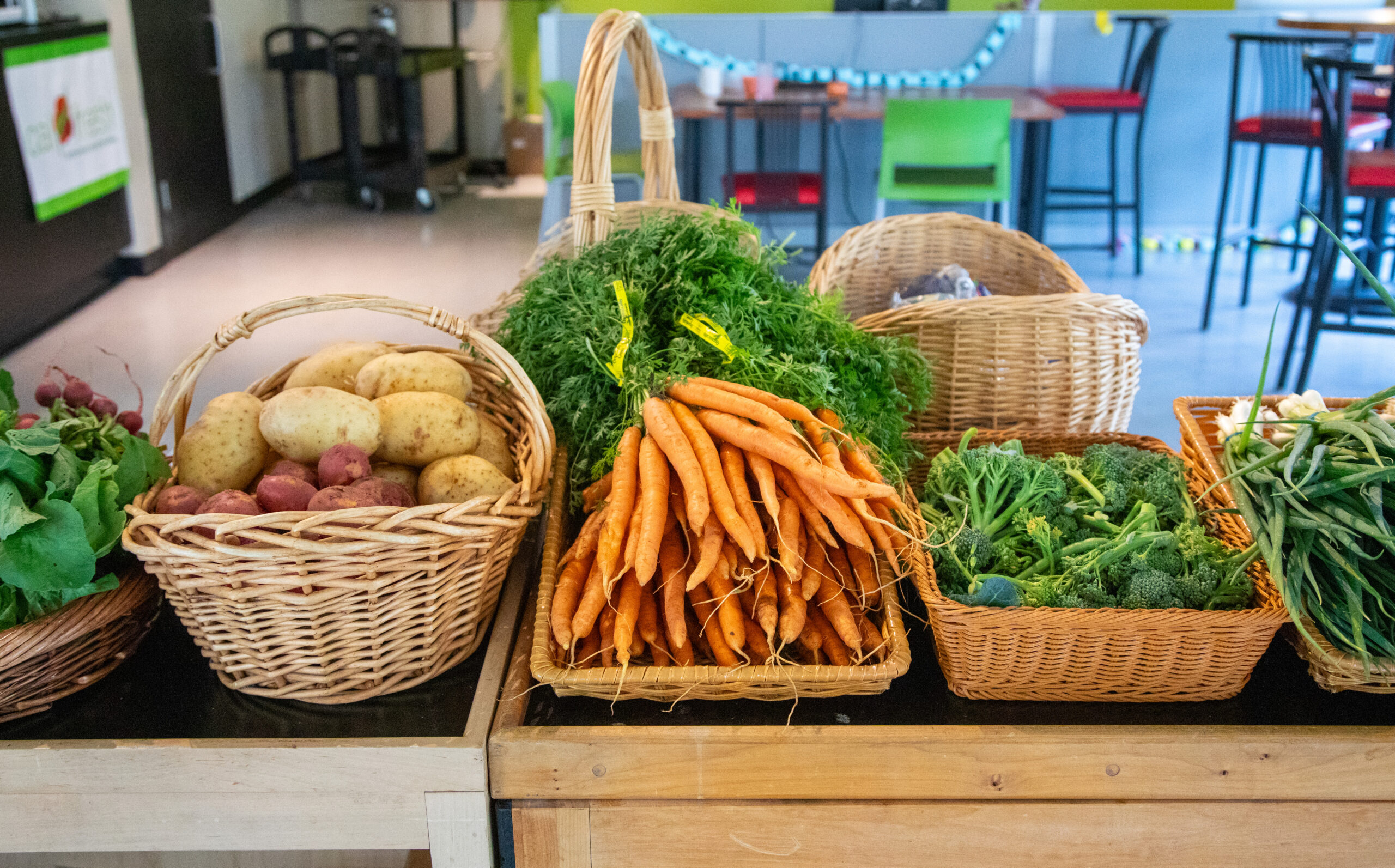 Baskets of fresh vegetables, including carrots, potatoes, broccoli, and green beans, are displayed on wooden tables. The setting appears to be an indoor market or produce stand.