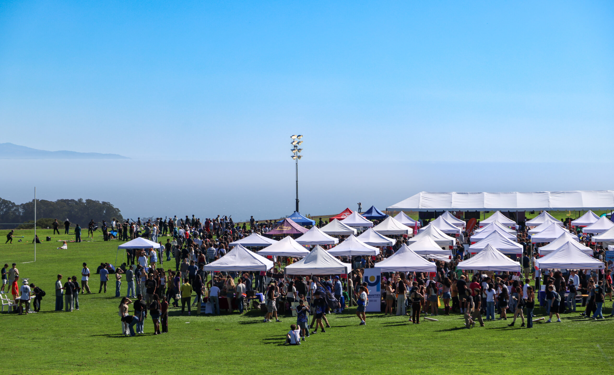 Students gather around rows of organization booths and tents on a large campus field overlooking the Monterey Bay.