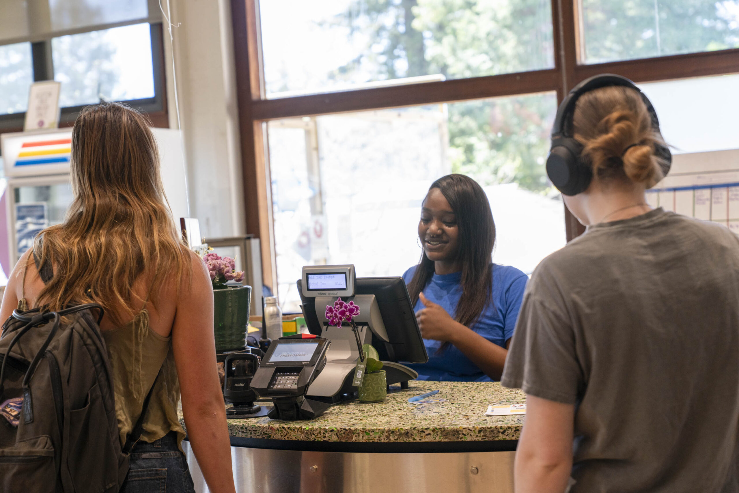 Two students stand at a campus dining counter while a student employee smiles and assists them at the register.