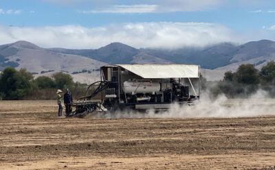 A steam injection machine billows steam as it moves across bare farm soil.