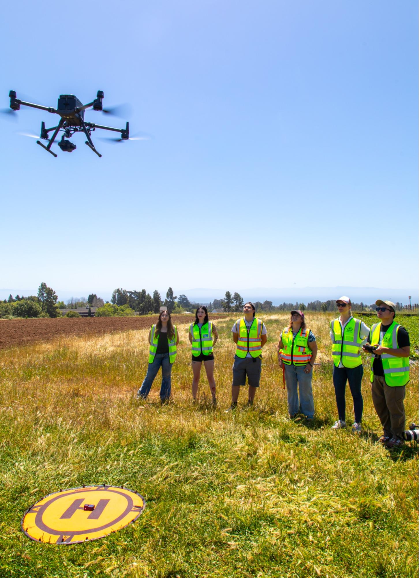 A group of six people in reflective vests observes a flying drone over a grassy field with a marked landing pad nearby. One person is holding a remote control, operating the drone.