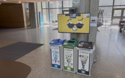 A waste station with bins for recycling, compost, and landfill sits in a bright, open lobby. Above the bins, a screen with a smiling face and sunglasses is mounted on a concrete pillar.